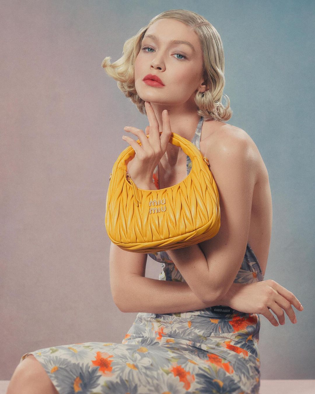 Gigi Hadid poses with a Miu Miu yellow leather handbag, wearing a floral print dress in a studio setting with soft, diffused lighting.