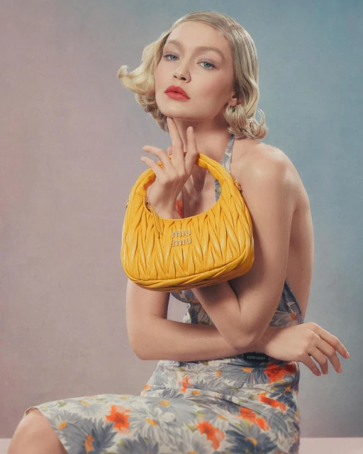 Gigi Hadid poses with a Miu Miu yellow leather handbag, wearing a floral print dress in a studio setting with soft, diffused lighting.