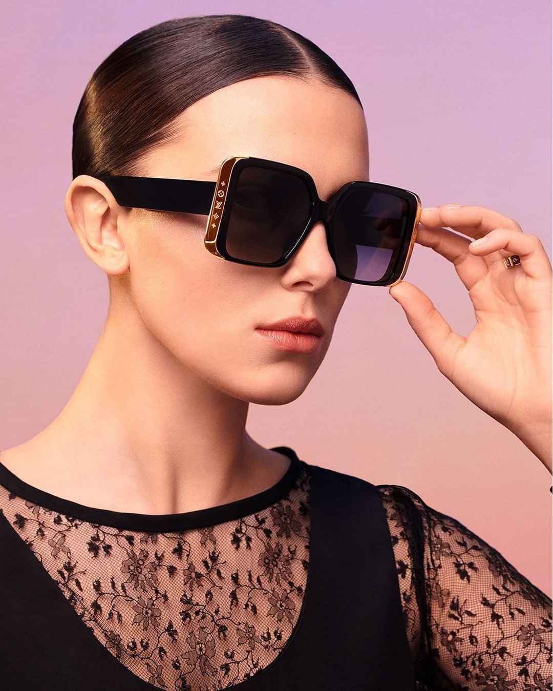 A woman with slicked-back hair wearing square sunglasses with embellishments poses against a purple gradient backdrop in a studio setting.