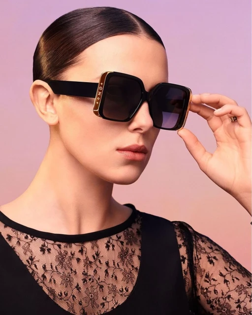 A woman with slicked-back hair wearing square sunglasses with embellishments poses against a purple gradient backdrop in a studio setting.