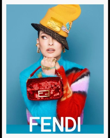 Fashion shot of adult female wearing a yellow hat and holding a red purse, posing in front of blue background with Fendi logo.