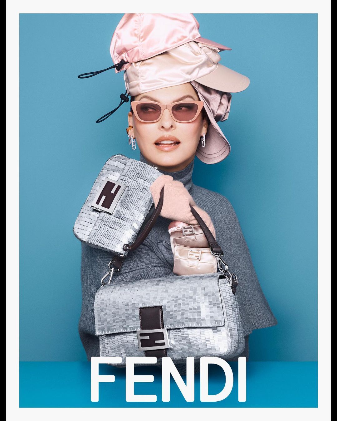 A model wearing a gray top and pink hat, holding Fendi bags, stands against a bright blue background, creating a striking fashion image.