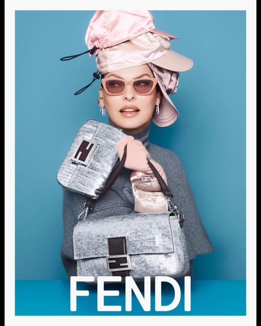 A model wearing a gray top and pink hat, holding Fendi bags, stands against a bright blue background, creating a striking fashion image.