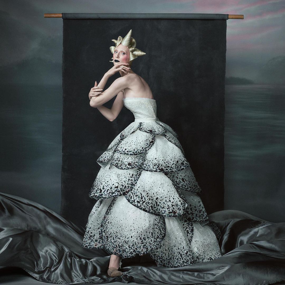 Elegant woman in tiered white and black dress with gold headpiece strikes pose against a textured backdrop, fashionably artistic.