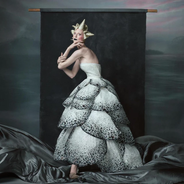 Elegant woman in tiered white and black dress with gold headpiece strikes pose against a textured backdrop, fashionably artistic.