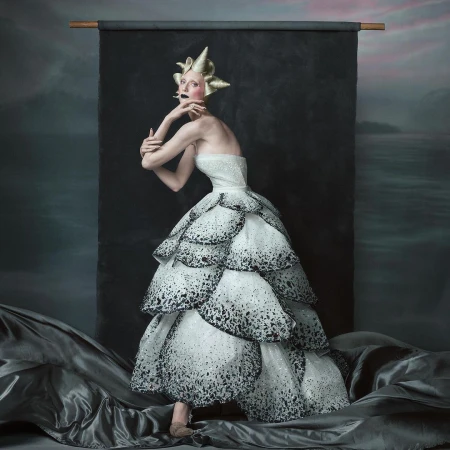 Elegant woman in tiered white and black dress with gold headpiece strikes pose against a textured backdrop, fashionably artistic.