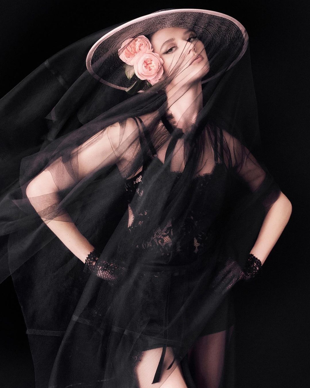 Woman in elegant black lace outfit, veil, and a hat with pink roses posing in a dark studio. Fashion, glamour, and portrait photography.