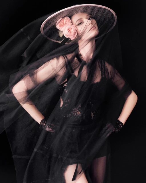 Woman in elegant black lace outfit, veil, and a hat with pink roses posing in a dark studio. Fashion, glamour, and portrait photography.