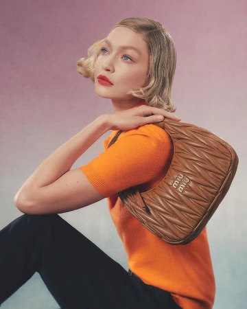 Blonde woman wearing an orange knit top holds a quilted brown leather handbag against a gradient backdrop