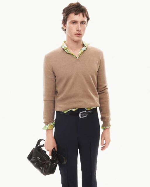 A fair-skinned adult male in a casual outfit poses against a white backdrop, holding a dark brown leather handbag. The composition focuses on fashion.