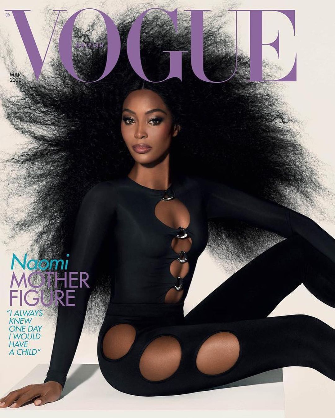 Naomi Campbell poses on a magazine cover in a black catsuit with circular cutouts, against a gradient white background. Big hair, close-up.
