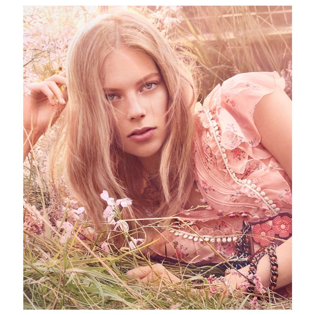Fashion shot of a blond woman lying in the grass with a pink floral top, bracelet, and pink flowers, with a warm color grading.