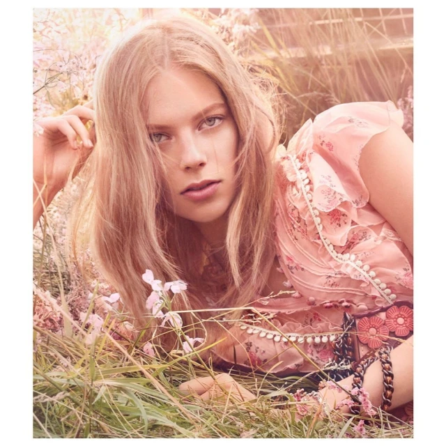 Fashion shot of a blond woman lying in the grass with a pink floral top, bracelet, and pink flowers, with a warm color grading.