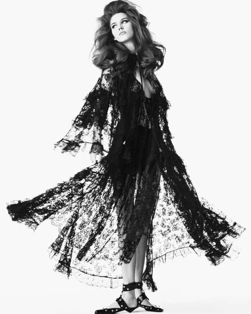 A woman with voluminous hair wearing a long, flowing black lace robe with studded sandals poses against a stark white background.