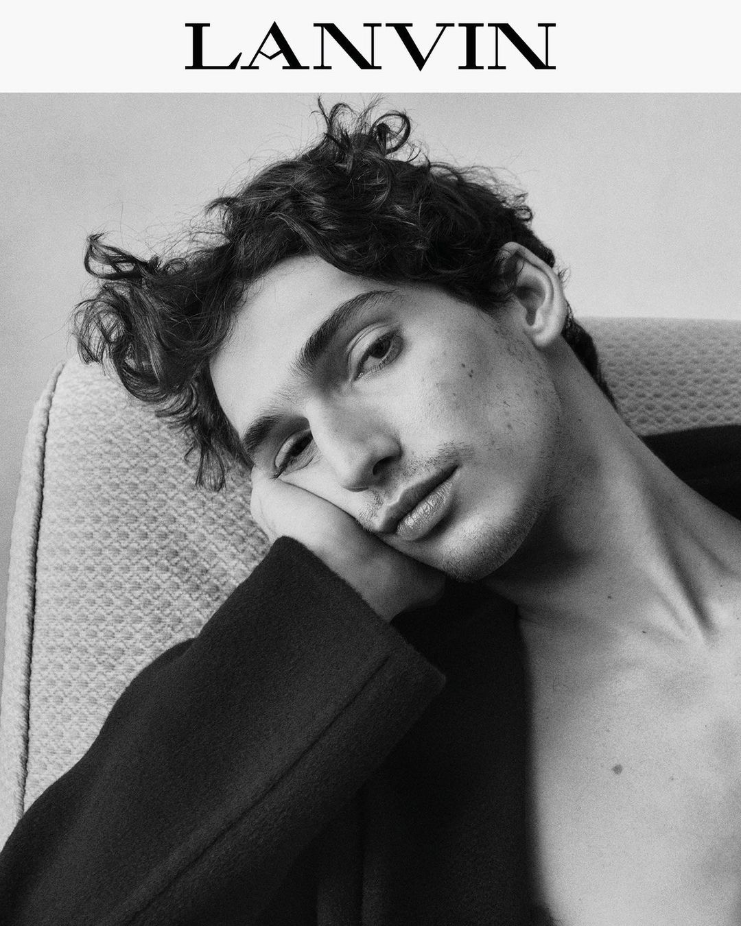 Monochromatic portrait of a young adult man with dark curly hair resting against an armrest with the Lanvin logo visible.