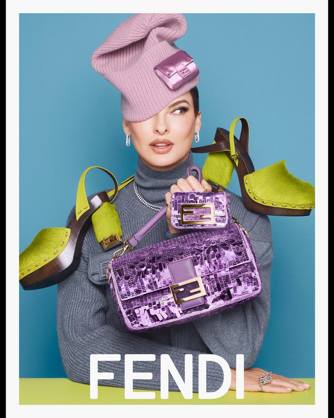 Woman wearing pink hat with handbag attached, grey turtleneck, holding a purple Fendi handbag. Green platform shoes and blue backdrop.
