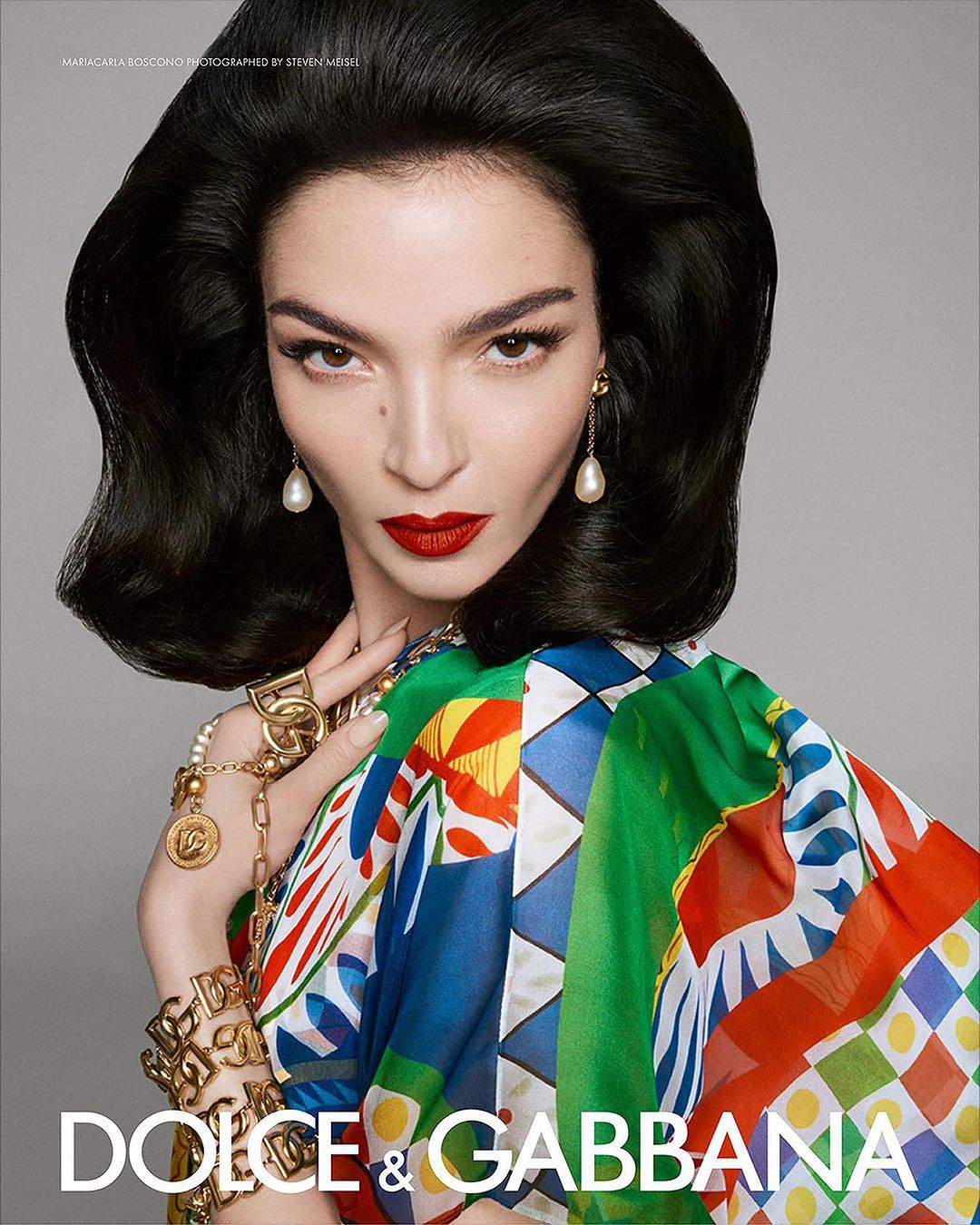 Glamorous woman with dark hair and red lips wearing colorful patterned shawl and gold jewelry posing in a fashion studio shot.