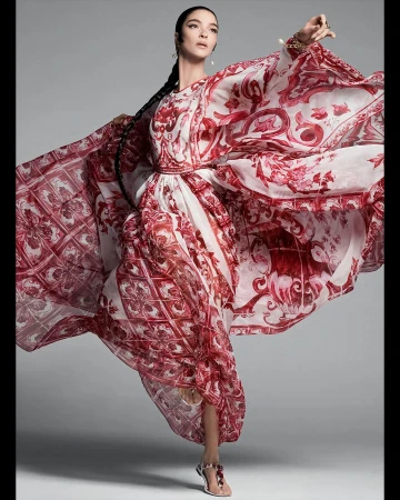 Model wearing a flowing red and white patterned dress with dramatic cape sleeves against a neutral grey studio backdrop.