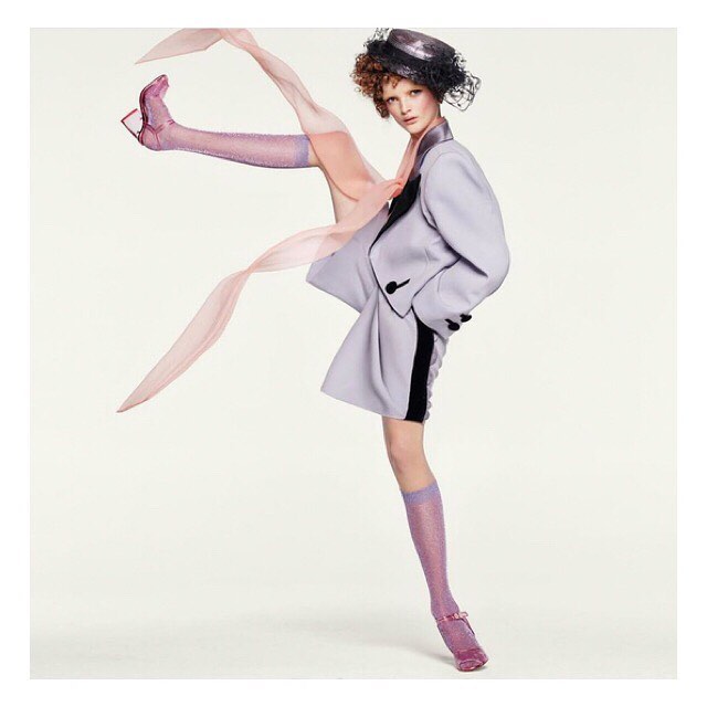 Fashion editorial shot: A woman in a pale purple suit and sparkly socks poses against a bright white backdrop with light pink accents.