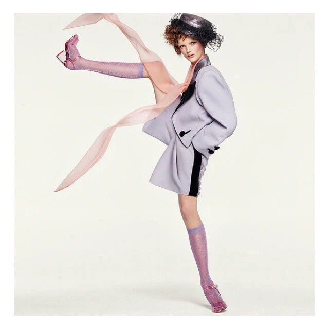 Fashion editorial shot: A woman in a pale purple suit and sparkly socks poses against a bright white backdrop with light pink accents.