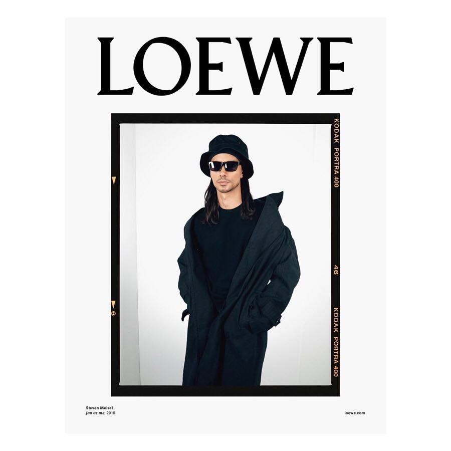 Fashion advertisement featuring a man in sunglasses, a bucket hat, and a long coat against a white background, framed by a black border with text.