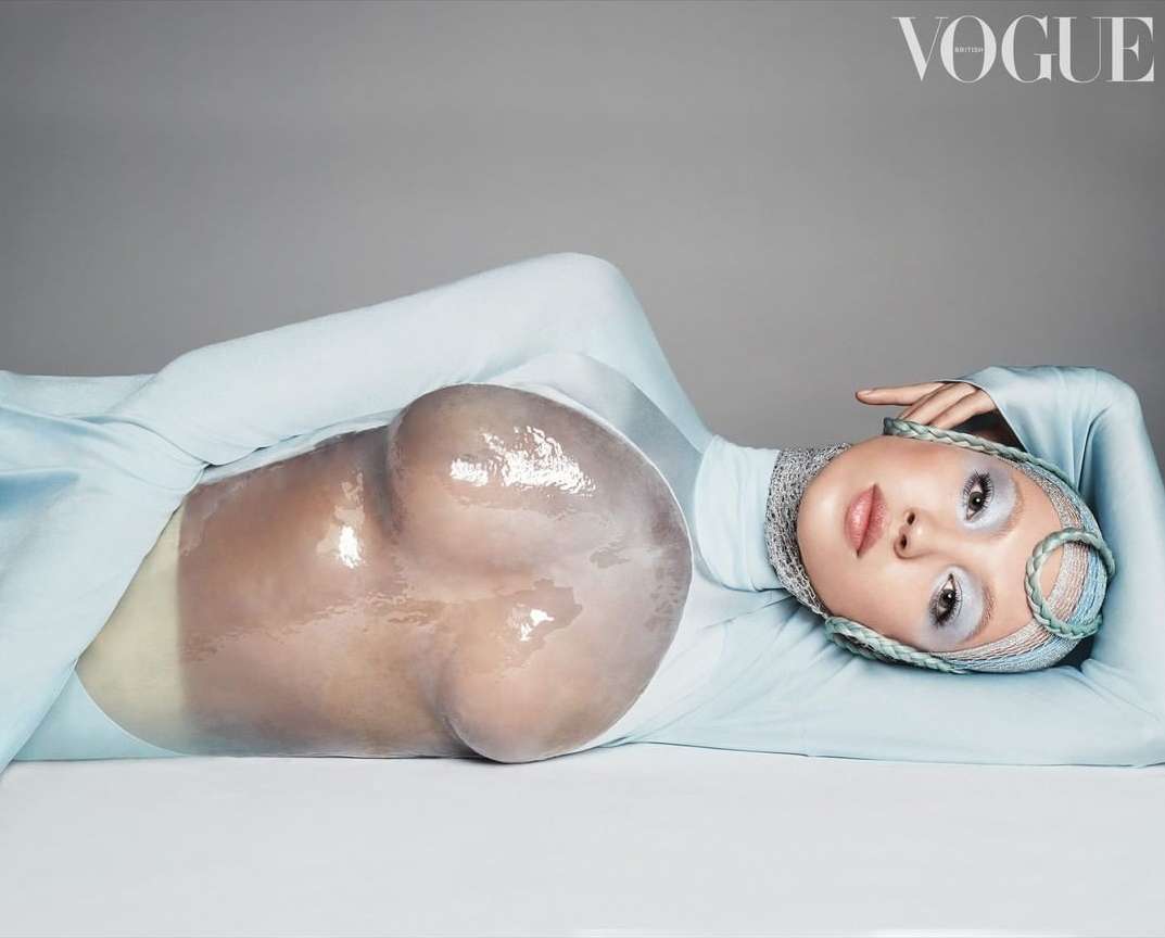 Fashion editorial shot of a model lying down wearing a light blue body suit and head covering with a plastic bubble over the abdomen.