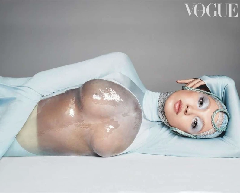 Fashion editorial shot of a model lying down wearing a light blue body suit and head covering with a plastic bubble over the abdomen.