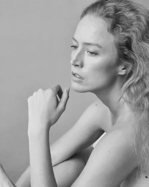 Black and white portrait of a woman with wavy hair. She is sitting with her hand near her face in an indoor photo studio.