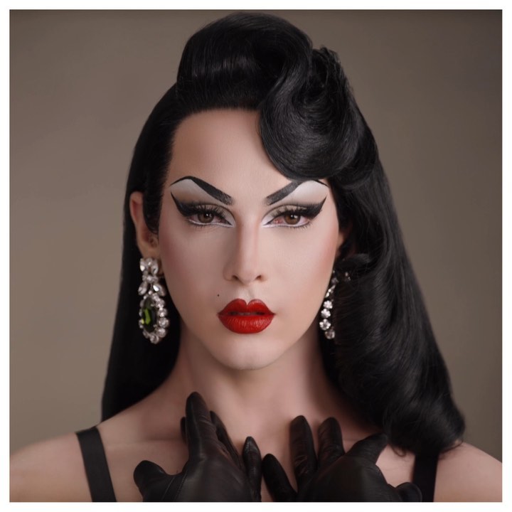 Portrait of a drag queen with dramatic makeup, black gloves, and vintage-style black hair against a neutral-toned background.