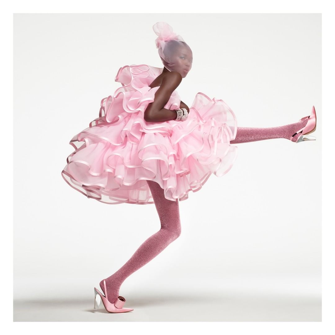 Fashionable Black model strikes a pose in a voluminous pink ruffled dress, matching tights, and heels against a clean white backdrop.