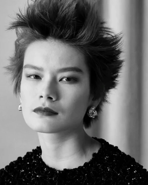 Close-up black-and-white portrait of a woman with spiky dark hair, dark lipstick, and statement earrings against a neutral backdrop.