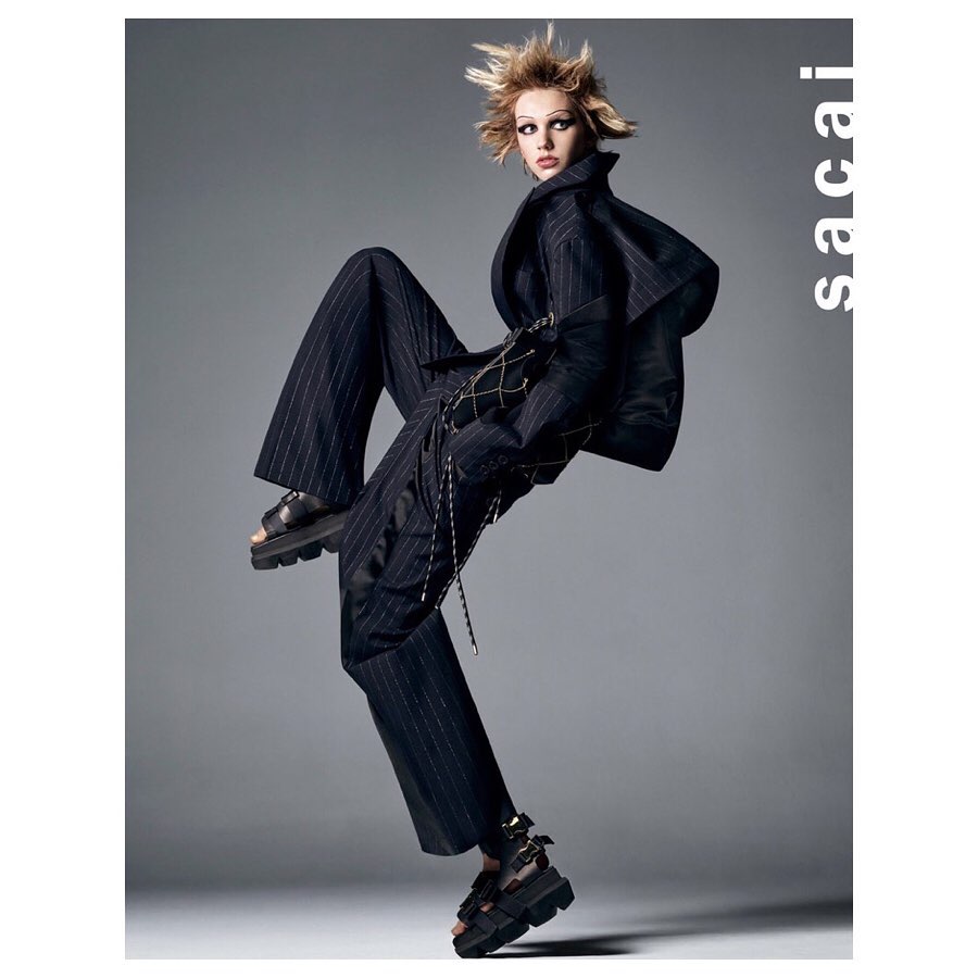 Fashion editorial shot of a blonde model in a pinstripe suit and black platform sandals, jumping in a grey photo studio for Sacai.
