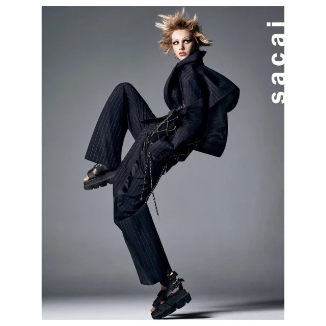 Fashion editorial shot of a blonde model in a pinstripe suit and black platform sandals, jumping in a grey photo studio for Sacai.
