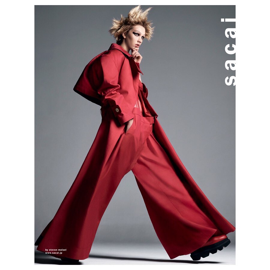 Fashion editorial shot: adult female model in a red coat and wide-leg pants strikes a pose against a grey backdrop with dramatic lighting.