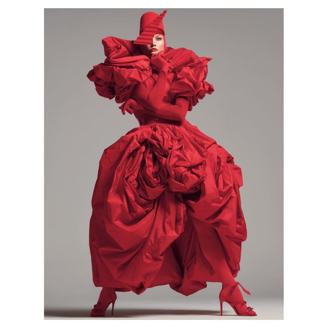 Fashion model in an oversized red gown, gloves, and headpiece poses in a studio with a grey background; full-length studio shot.