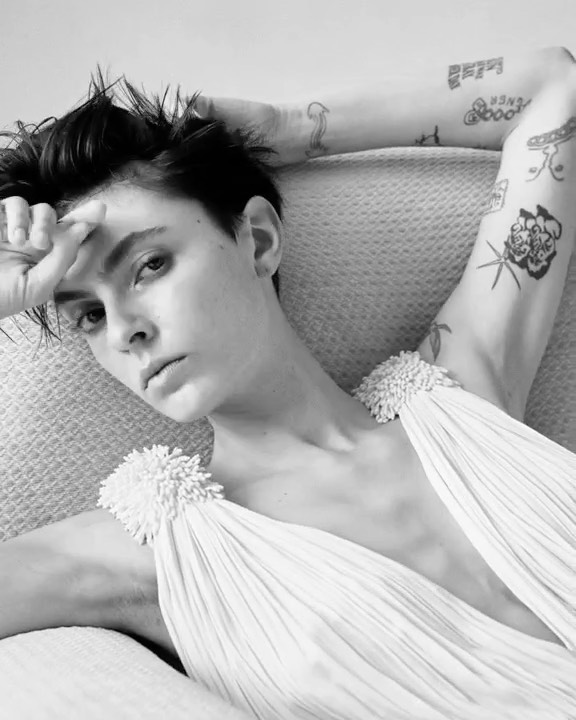 Monochrome portrait of a woman with tattoos reclining in a light-colored dress, posing indoors with a hand touching her forehead.