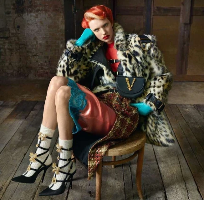 Woman seated in a leopard print coat, red top, patterned skirt, black heels with butterfly details, and turquoise gloves in a room.