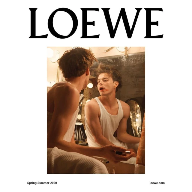 A young adult male in a tank top applying lipstick while looking into a mirror with vanity lighting and the Loewe logo above.