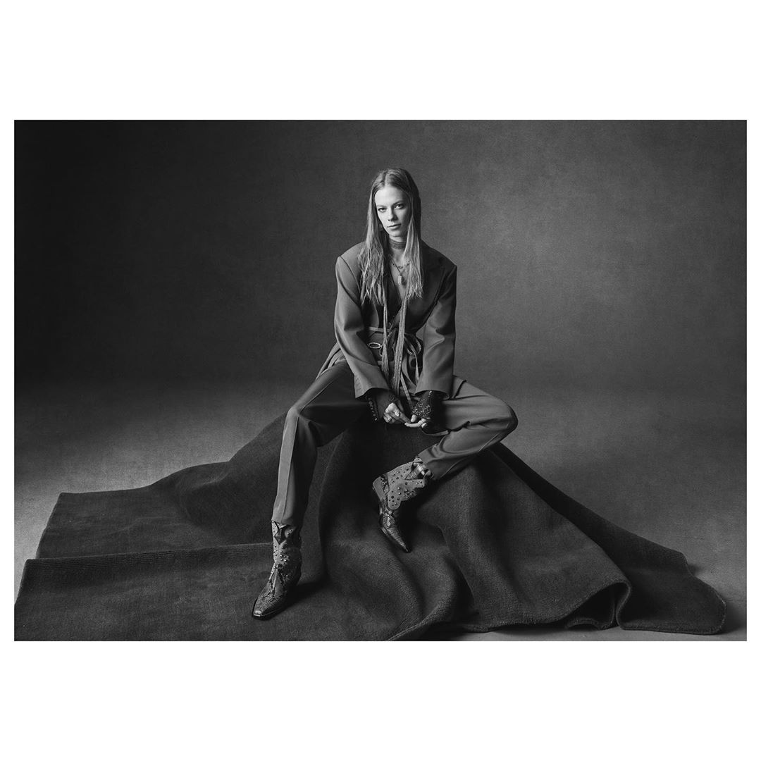 Monochrome fashion portrait of a poised woman in a pantsuit and boots, seated on fabric against a muted backdrop in a photo studio.