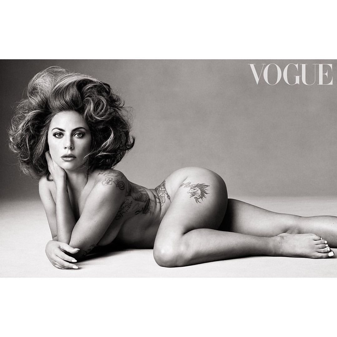 Monochrome nude portrait of a woman with curly hair reclining against a grey background, tattoo art visible on her body, Vogue logo.