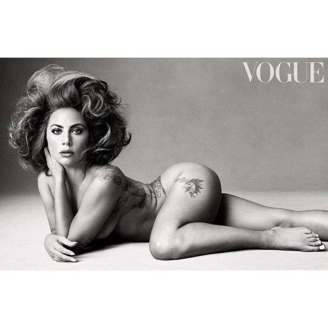 Monochrome nude portrait of a woman with curly hair reclining against a grey background, tattoo art visible on her body, Vogue logo.