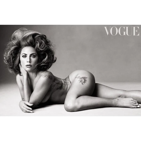 Monochrome nude portrait of a woman with curly hair reclining against a grey background, tattoo art visible on her body, Vogue logo.