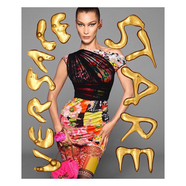 Bella Hadid poses in a colorful floral dress with gold abstract shapes and a pink purse against a grey backdrop in a fashion editorial shot.