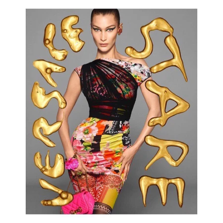 Bella Hadid poses in a colorful floral dress with gold abstract shapes and a pink purse against a grey backdrop in a fashion editorial shot.