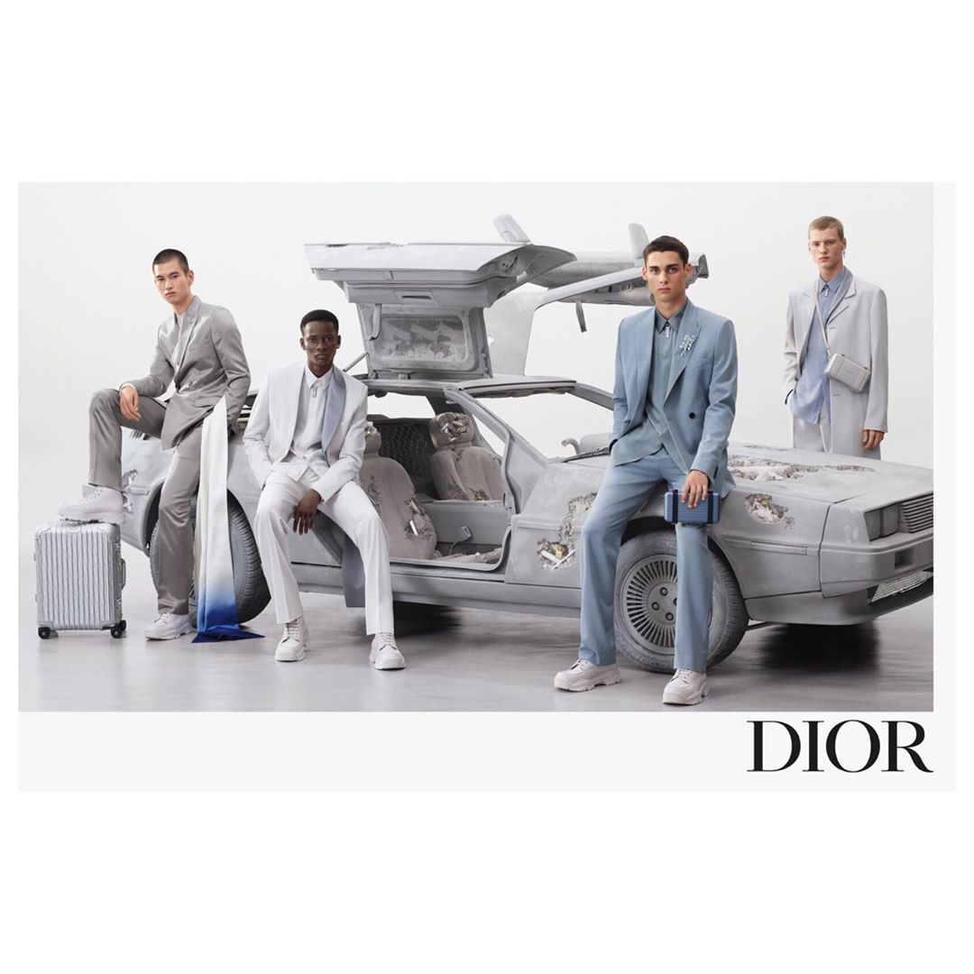 Four adult male models pose with a weathered DeLorean automobile against a plain background, highlighting fashion and automotive elements.