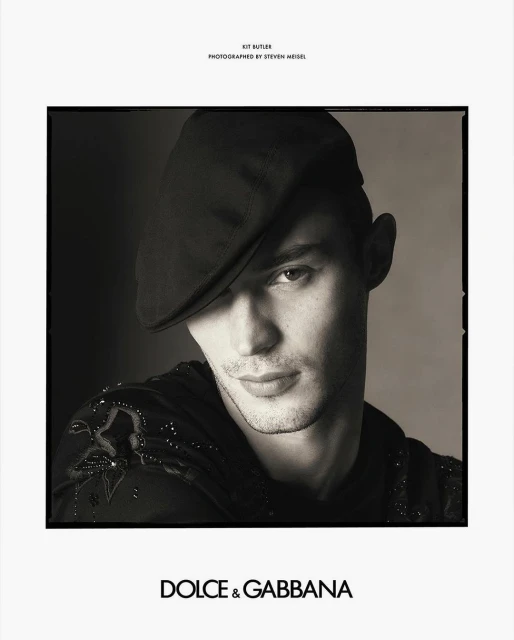 Monochrome portrait of a man in a newsboy cap and embellished top against a dark backdrop, promoting Dolce & Gabbana.