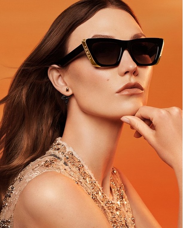 Elegant woman wearing stylish black sunglasses with gold accents, posing in a close-up studio shot against an orange background.