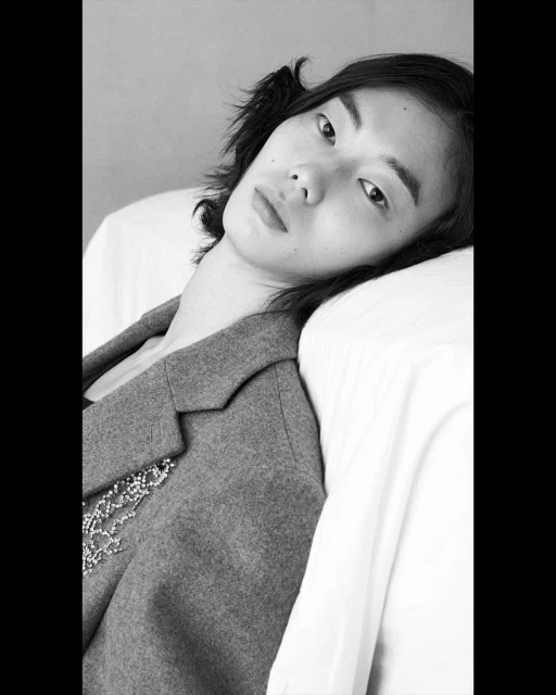 Monochrome portrait of a young adult posing in a blazer, leaning against a white cushion. Black and white tones give the image a timeless feel.