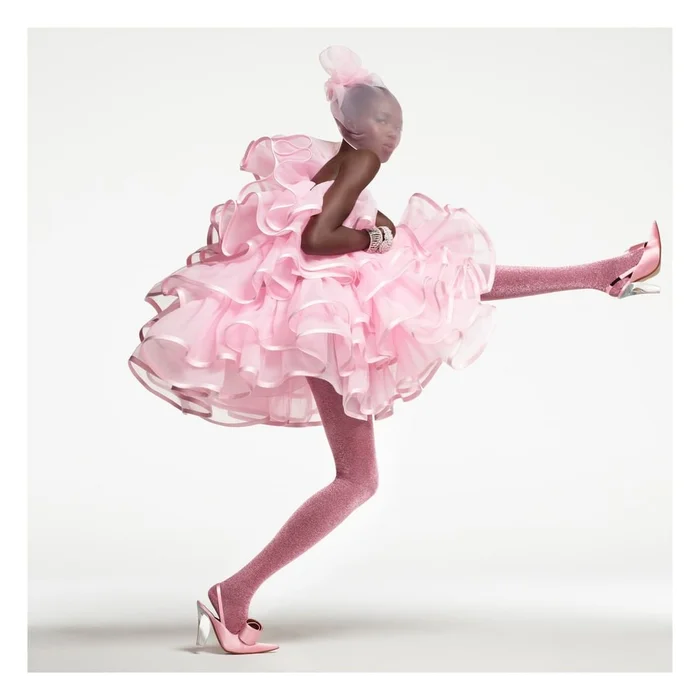 Model in voluminous pink ruffled dress and matching sheer veil kicking leg out against white studio background