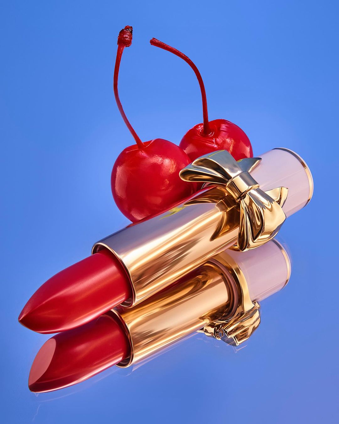 Red lipstick tube with gold bow, reflective surface, and cherries against a blue background in a beauty and fashion product shot.